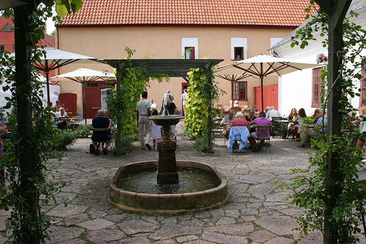 På innergården för kafét finns en fontän framför en pergola med växter. Bredvid pergolan ätande eller fikande gäster runt bord.